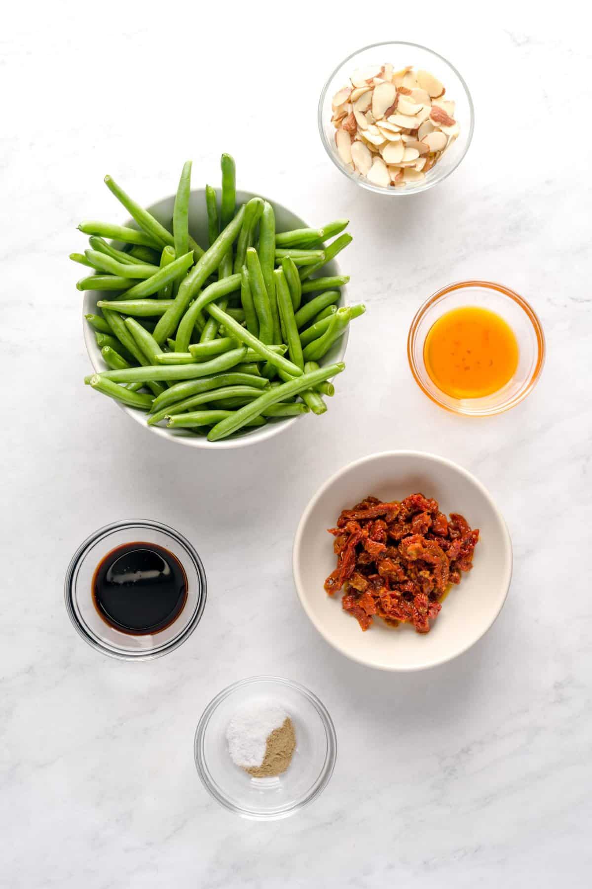 Overhead shot of ingredients for Italian green beans, such as sliced sun-dried tomatoes, balsamic vinegar, extra virgin olive oil, sliced almonds, salt, pepper and fresh green beans.