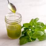 Spoon pouring basil olive oil into a jar while next to fresh basil.