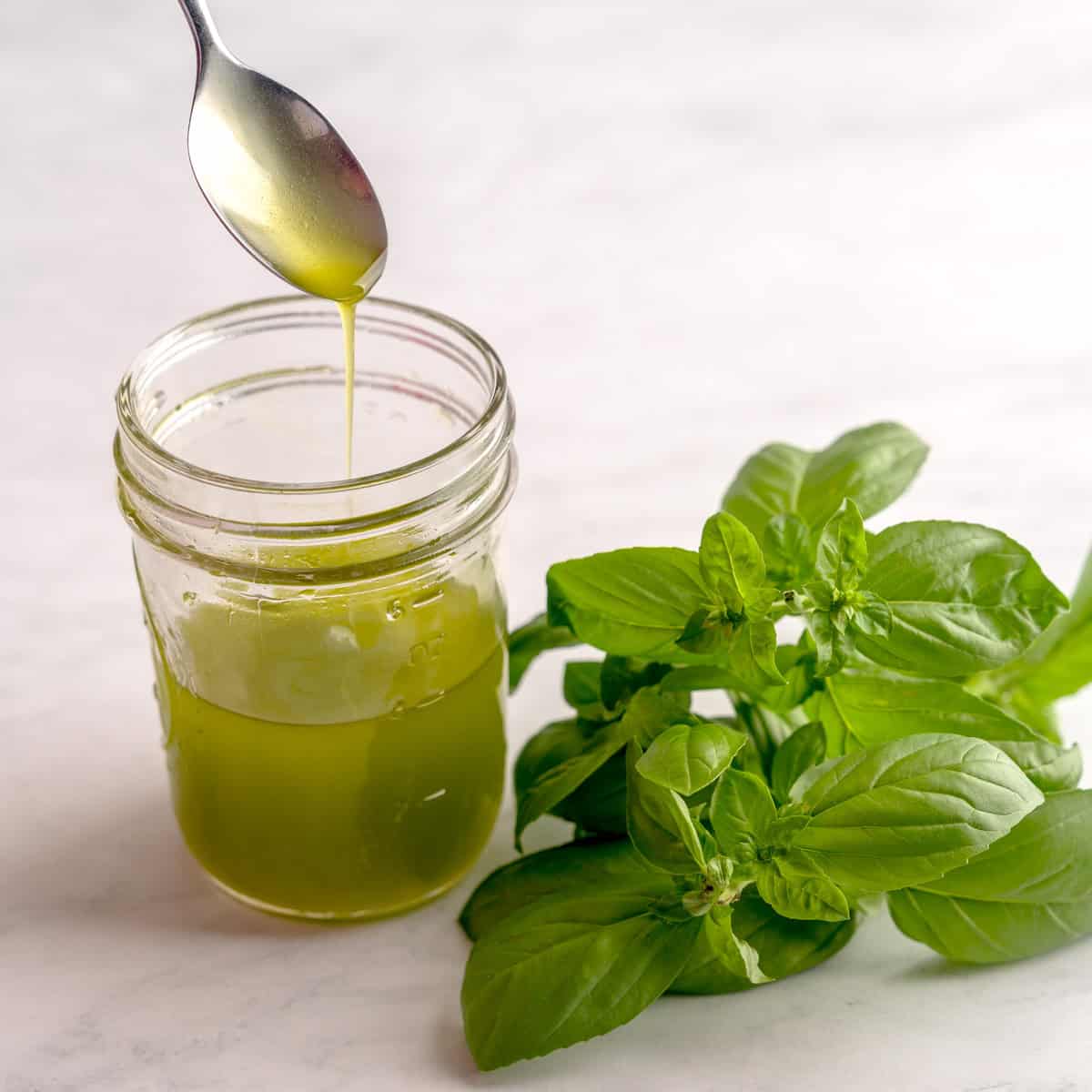 Jar of homemade basil-infused olive oil with fresh basil and a spoon drizzling oil on a white surfac