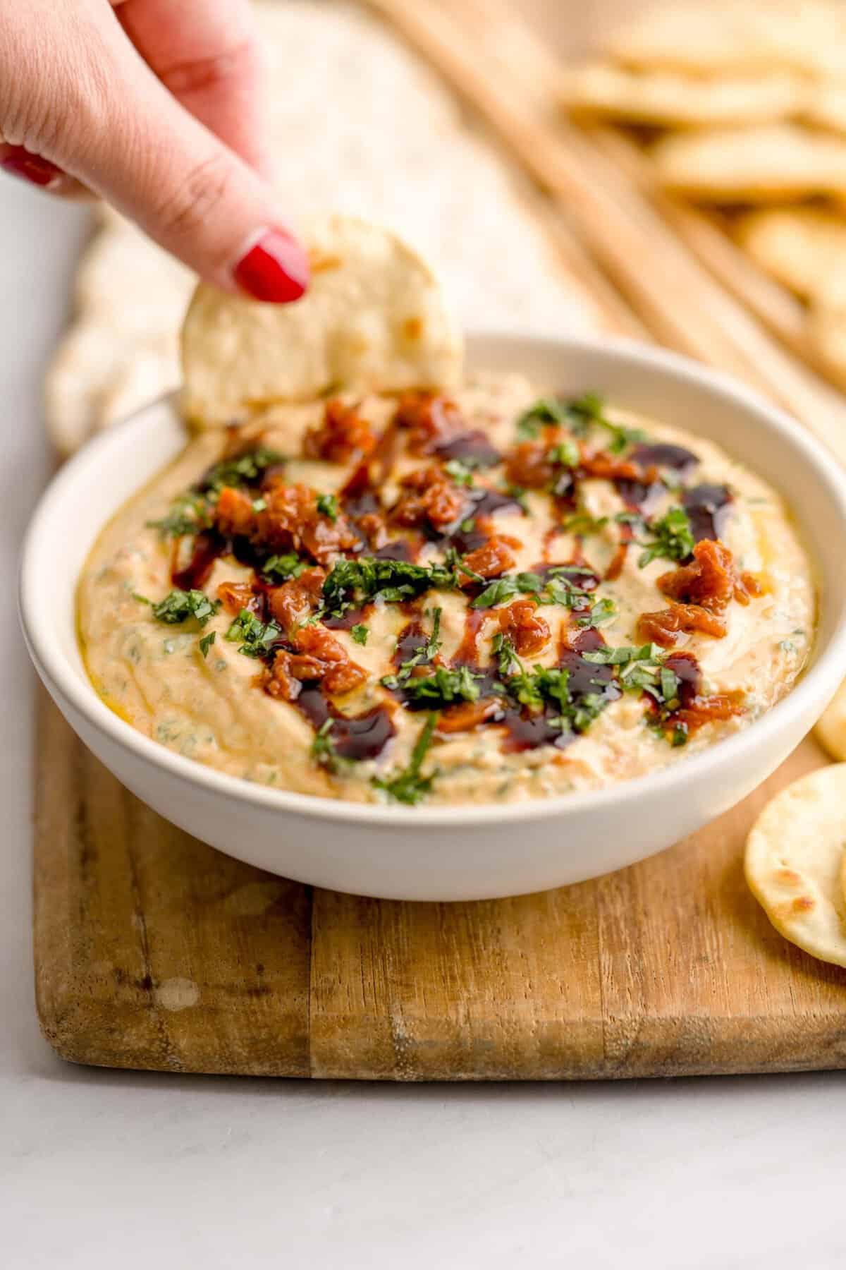 Dipping a pita bread cracker into a bowl of whipped ricotta dip with sun-dried tomatoes and basil.