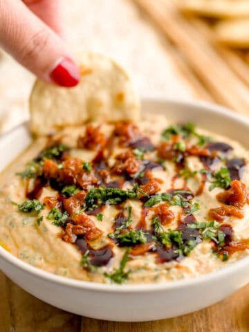 Dipping a pita bread cracker into a bowl of whipped ricotta dip with sun-dried tomatoes and basil.