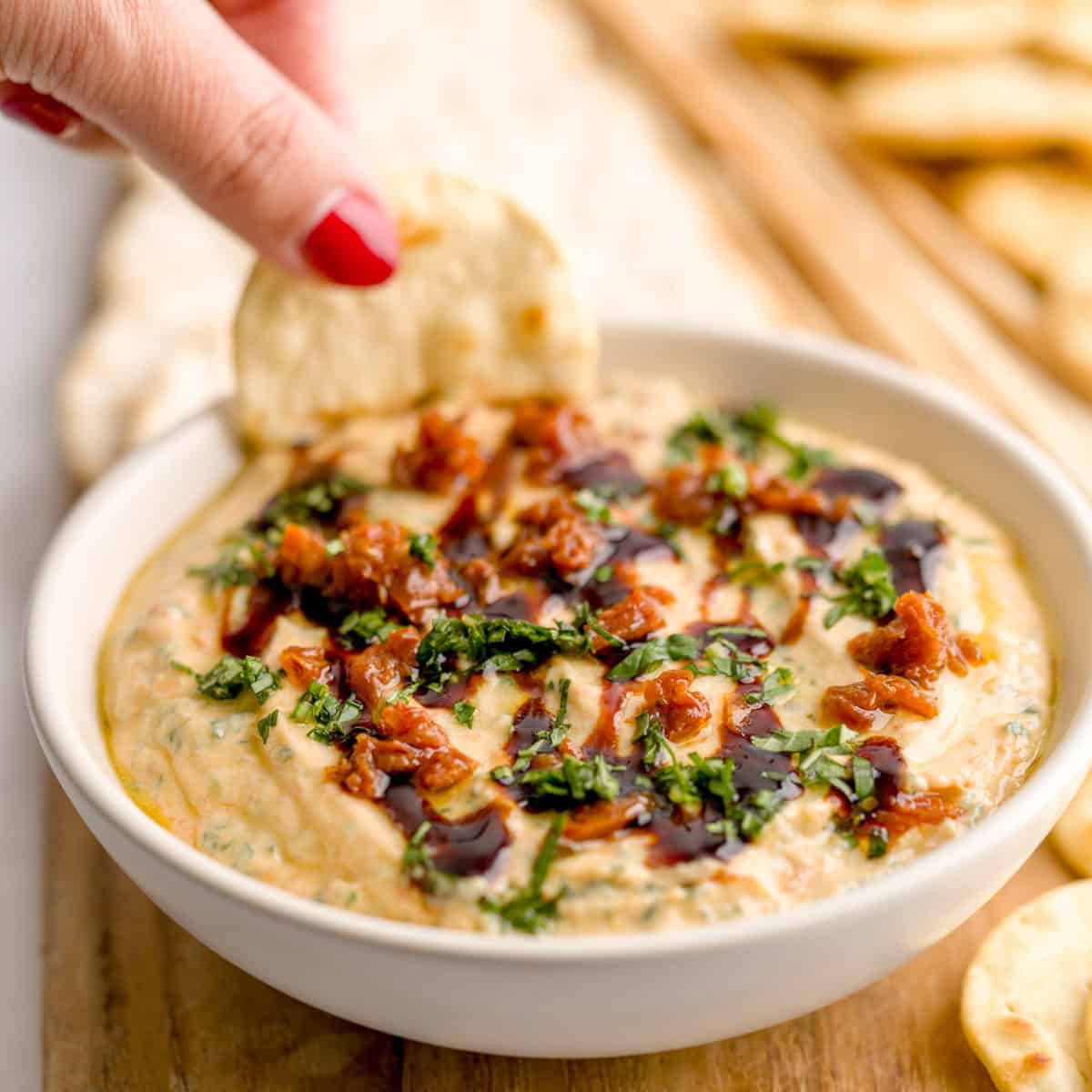 A hand dips a round pita chip into a bowl of whipped ricotta dip, topped with chopped sun-dried toma