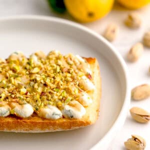 Closeup shot of basil-lemon ricotta toast surrounded by lemons and pistachios.