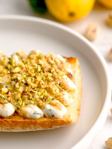 Closeup shot of basil-lemon ricotta toast surrounded by lemons and pistachios.