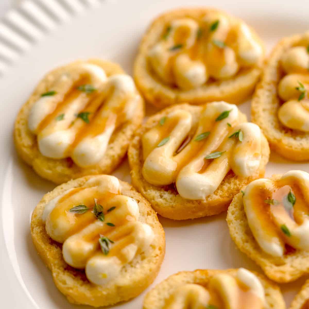 Crostini with whipped ricotta, truffle honey drizzle, and thyme on a white plate.