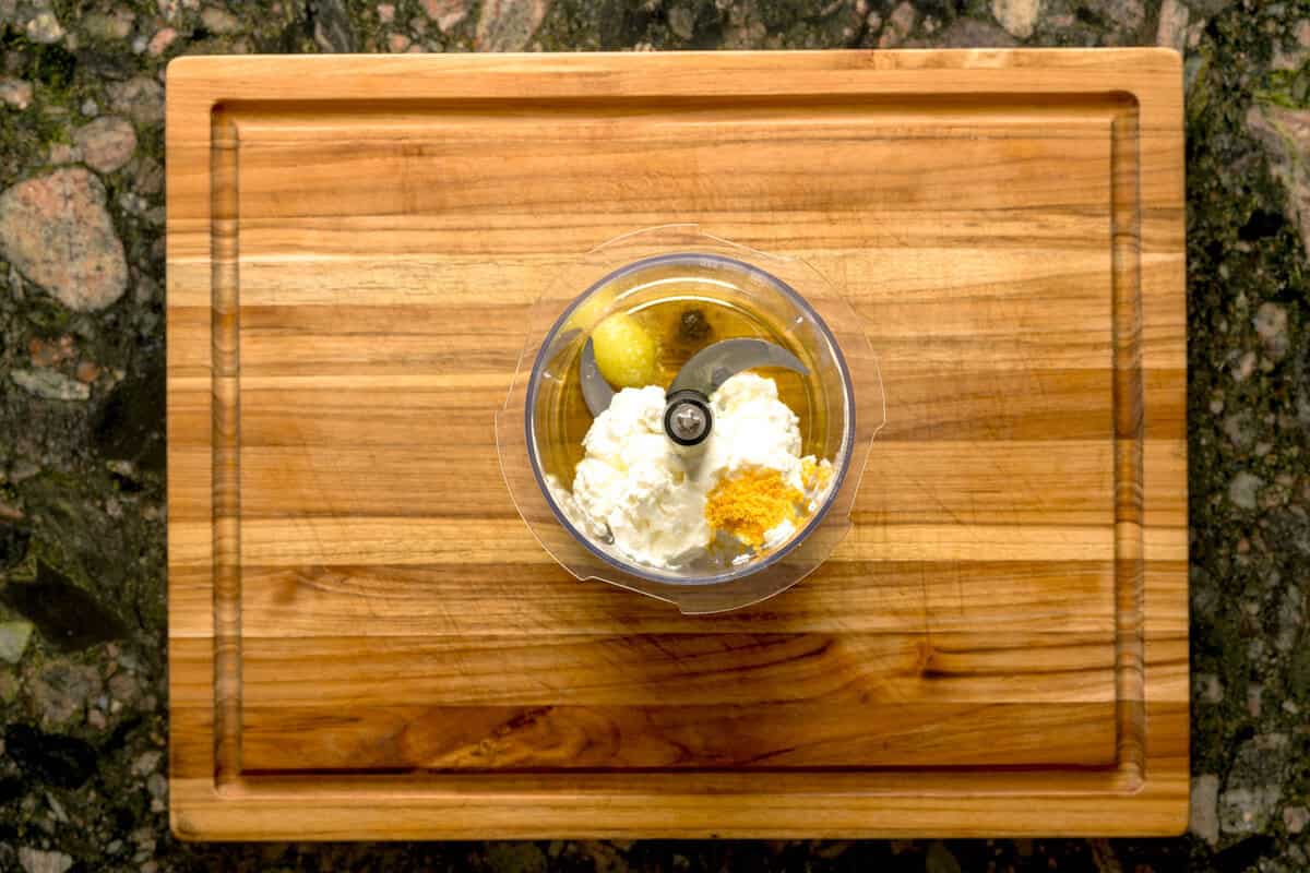 Overhead shot of food processor with ingredients to make whipped ricotta, like oil, ricotta and lemon zest.