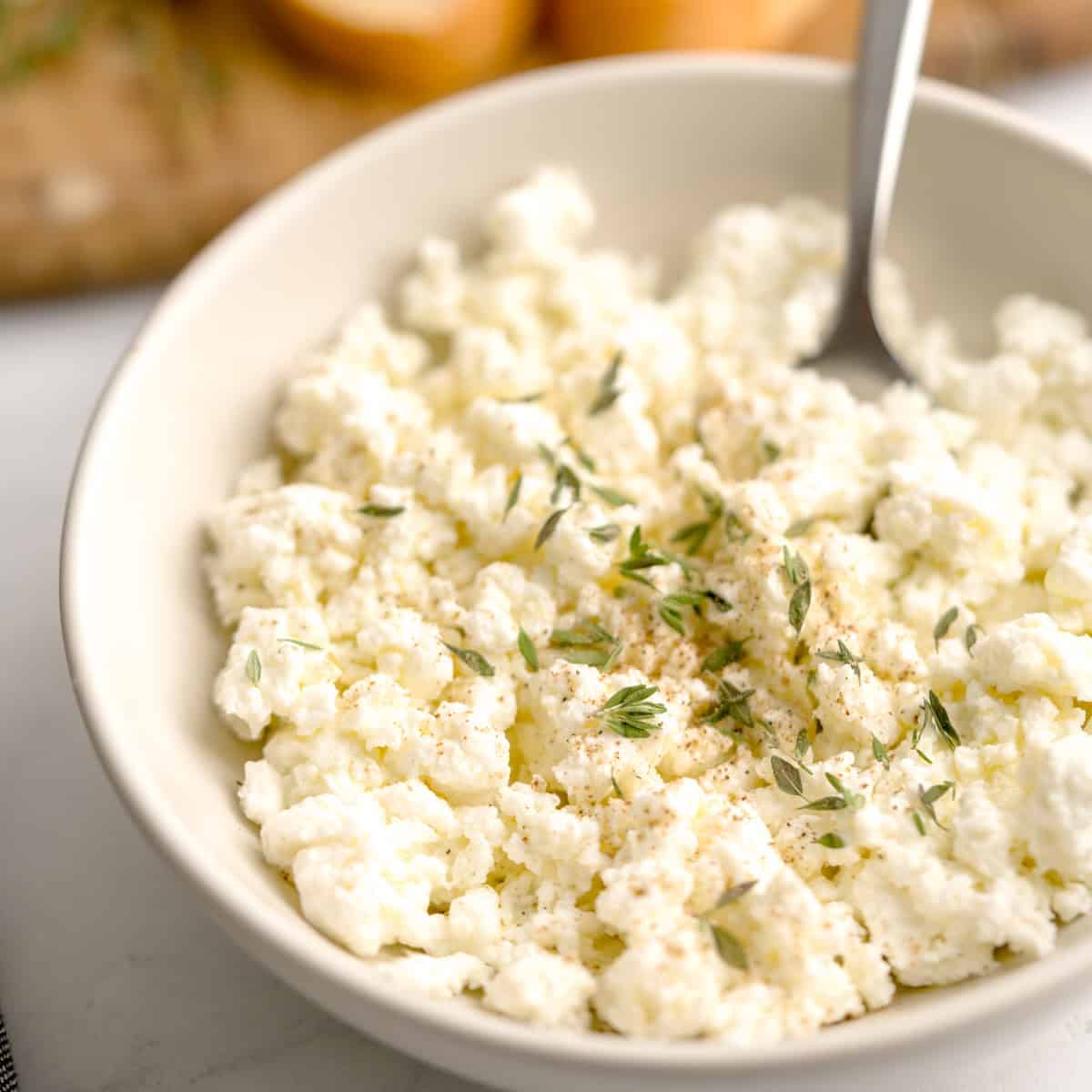 Bowl of homemade ricotta cheese topped with fresh herbs, served with a spoon on a white surface.