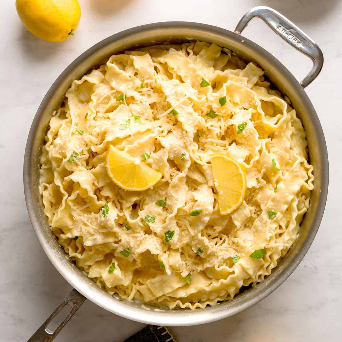 Creamy lemon pasta in a stainless steel pan topped with lemon wedges, grated cheese, and fresh herbs