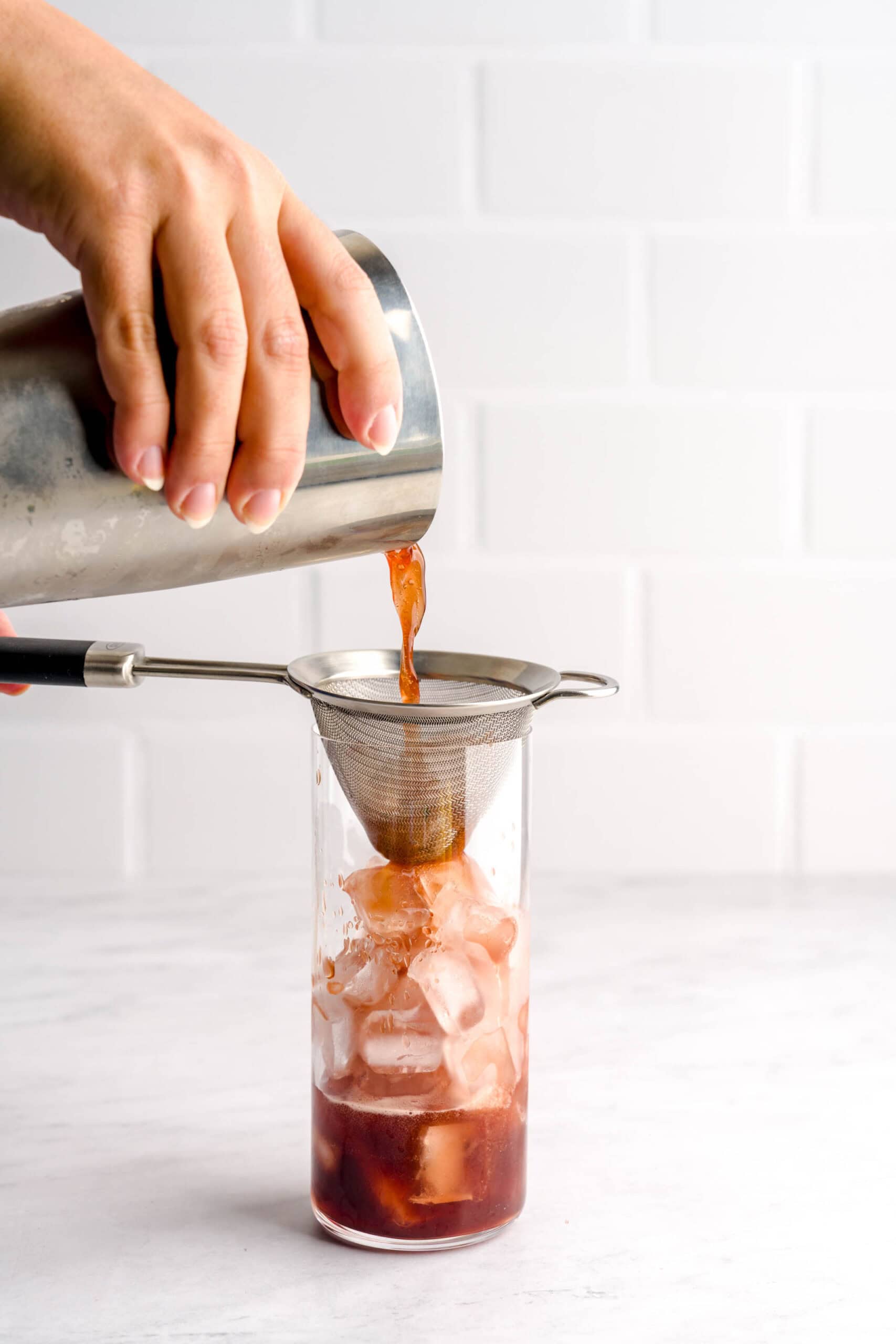 Straining Italian-inspired basil pomegranate cocktail into a tall glass with ice.